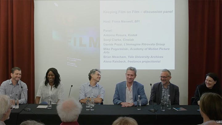 Six people--four men and two women--sit at a long table with black cloth. Projected behind them on screen reads "Keeping Film on Film" discussion panel, Host: Fiona Maxwell, BFI and names of panelists