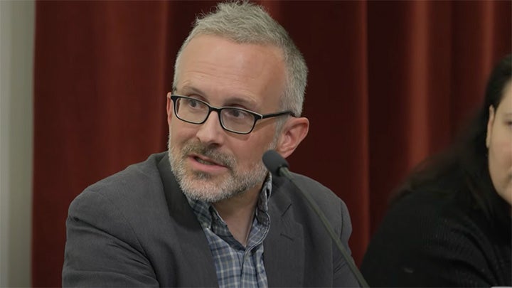 Closeup of man with short grey hair and beard and black rim glasses, turning head to right while speaking at microphone. He wears grey jacket and blue and gray plaid shirt open at collar.