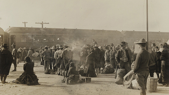 1914 sepia-tone photo showing crowds of people with suitcases, in a dusty setting, some seated some standing, with two train cars in the near distance