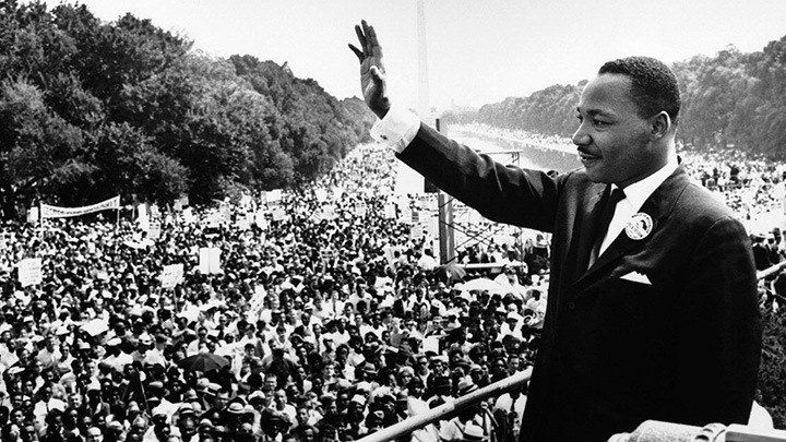 Black and white photo of man in suit and tie with arm raised to greet the hundreds of people in crowd below
