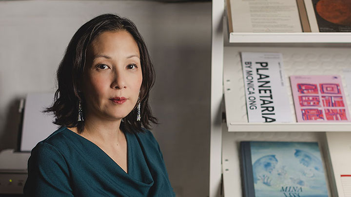 Woman with short dark hair and dark green blouse and long earrings looks at camera. To her left are shelves that display artwork. One reads "Planetaria by Monica Ong"