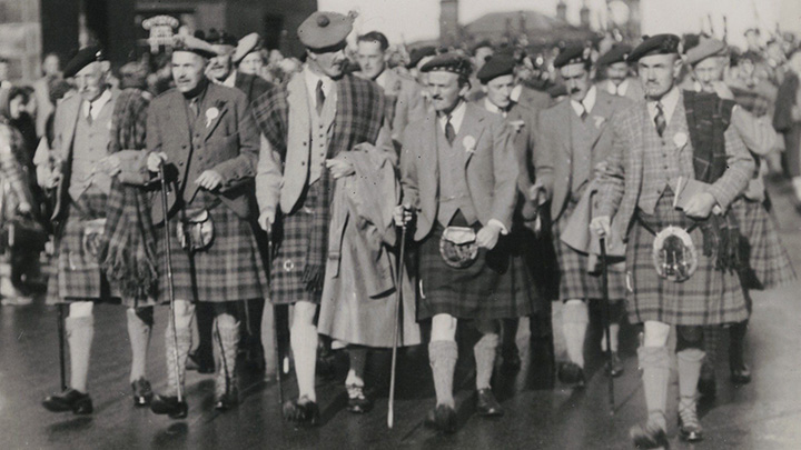 Black and white 1914 photo of large crowd of men in Highland dress marching with walking sticks