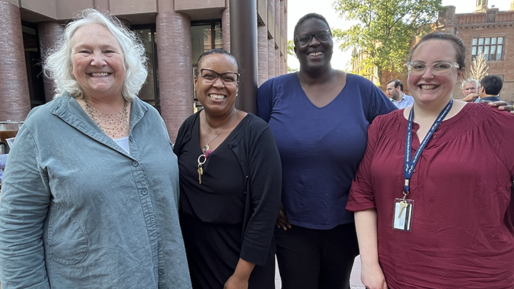 Four smiling women post: one with white hair and blue shirt, one with short black hair, glasses and black top, one with short black hair, glasses, and dark blue top, and one with glasses and red shirt