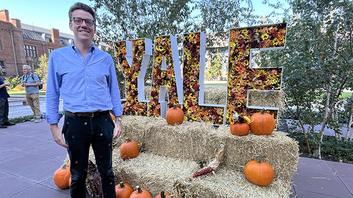 Man with blue shirt and dark green pants and glasses stands in front of display of haystacks and pumpkins with YALE sign filled with yellow and gold flowers