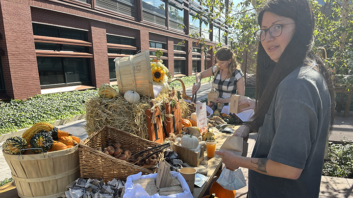 Woman with long dark hair and dark rim glasses looks at camera and stands in front of table full of colorful gourds, baskets, and doughnuts. Woman in background reaches for doughnuts with tongs