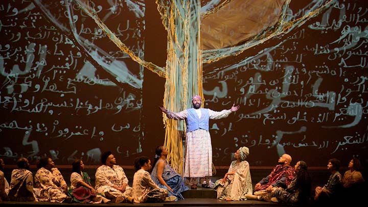 Stage set with arabic script projected in white on black background. Man in Arabic dress stands with arms outstretched in front of animated image of a blue and yellow tree. Eleven people in long garments sit at his feet, six to his right and five to his left.