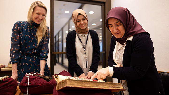 Three women gather around open book. A blond woman wearing dark blue dress stands next to a shorter woman wearing white shirt and black sweater and beige hijab. Woman, wearing black sweater and white shirt and maroon hijab, at far right points to open page in the book. 