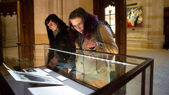 Two women peer into a lit display case. One has long black hair and a black jacket. The one to her left has shoulder-length hair and glasses, wears a beige coat and points to an object in the case.