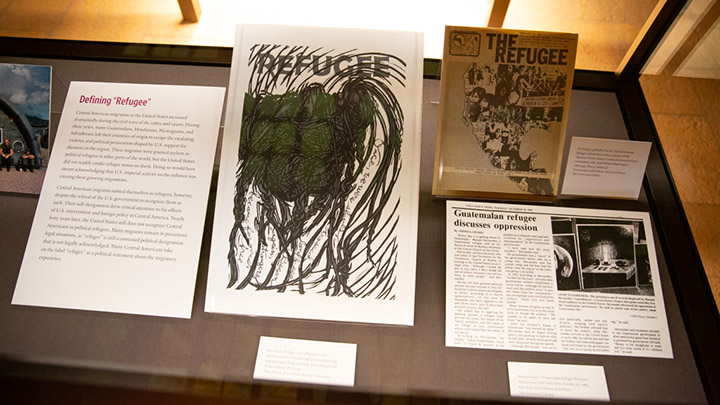 Closeup view of interior of display case, showing three documents in black and white. The poster reads "Refugee." The title on another page reads "The Refugee" and shows a photograph of people protesting. To the left is a typed descriptive label.