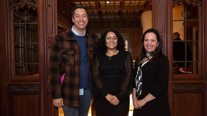 A tall man and two women pose, smiling for the camera. The man has a brown and black jacket over a blue sweater and white shirt. The woman at center has black hair, glasses, and a black dress. The woman at far right has long dark hair, a dark dress, and a long black, white, and brown scarf.