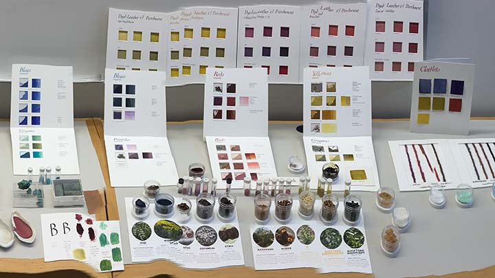 Table layout of 10 white cards that show swatches of color, ranging from blues, yellows, purple. In front of the standing cards are open plastic cylinders containing ground pigments in a range of colors, used to make paints. At lower left is a white paper with some brush strokes of sample colors--four green, four purple, and one yellow