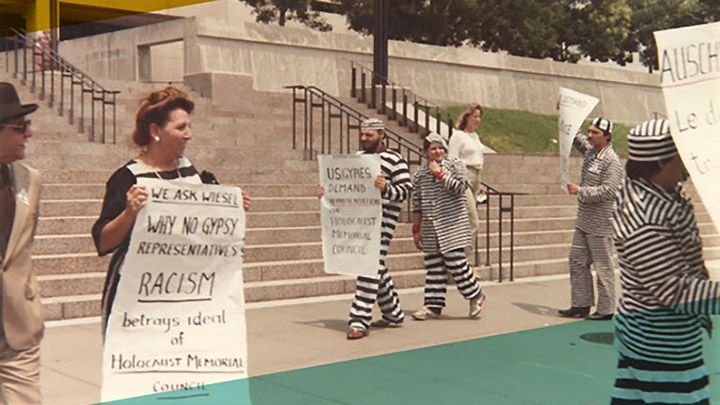 Four protesters, two carrying large placards. One reads "Why no Gypsy representations, Racism betrays idea of Holocaust Memorial." Two are dressed in prisoner garb