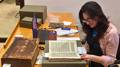Seated woman with dark hair and glasses peruses old book resting in a mount on the table in front of her. Other special collection materials, boxes, papers an an open laptop are spread across the tabletop.