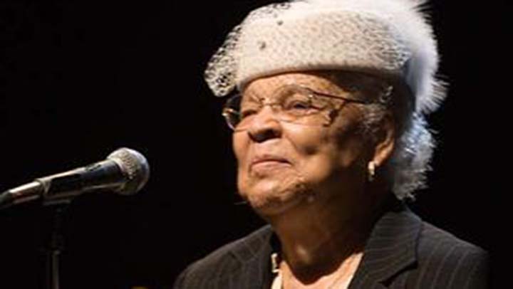 Elderly woman wearing a beige pillbox hat with veil stands before microphone smiling. She wears wire-rimmed glasses and a black jacket.