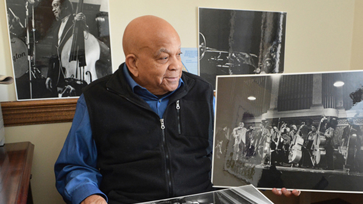 Bald man wearing blue shirt and dark blue zippered vest sits looking to side, black and white photograph of jazz musicians and Duke Ellington sit in front of him, and photograph of man when younger holding acoustic bass is behind him on wall, next to another photo showing a man playing a trumpet
