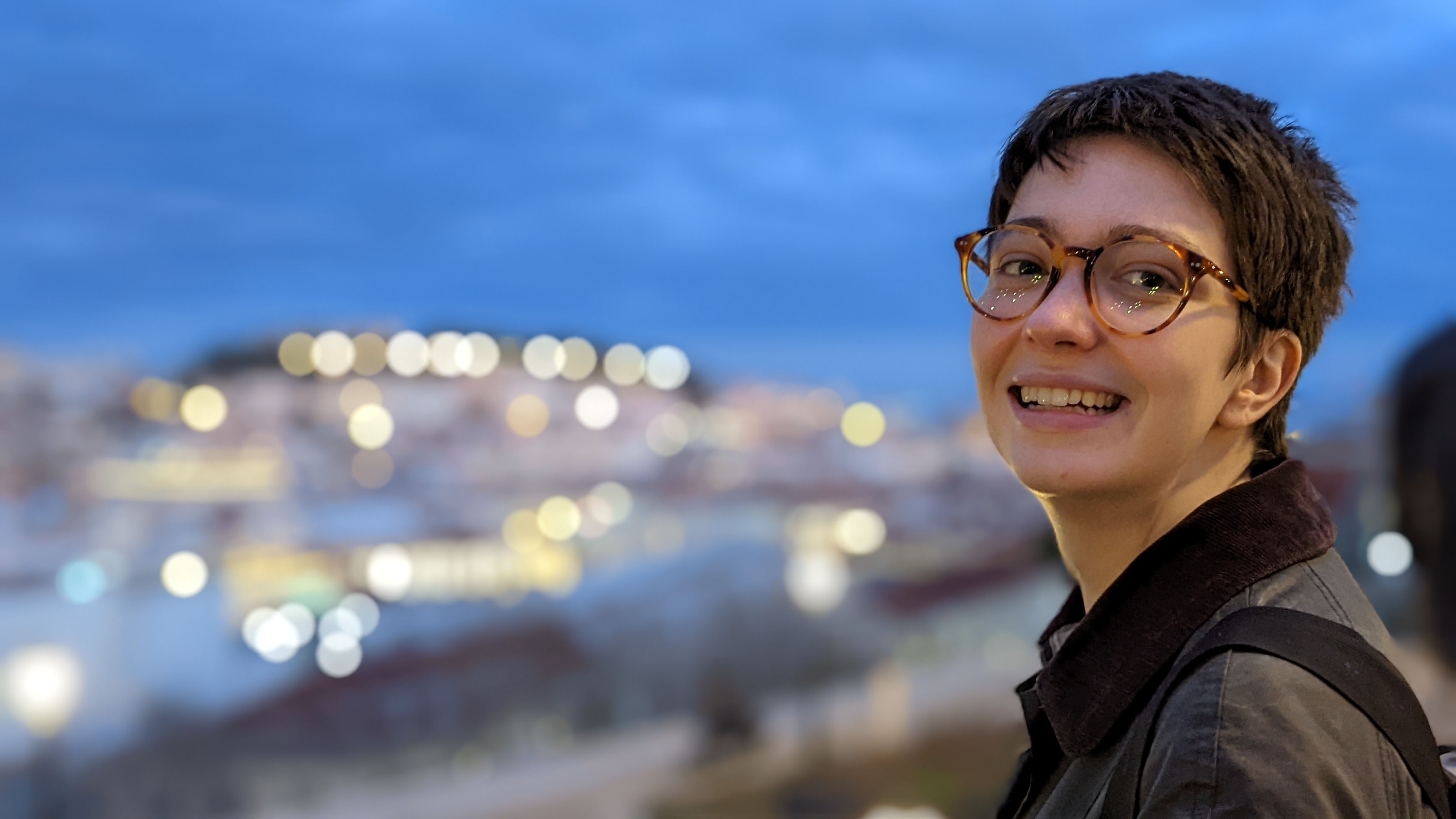 Smiling female student with shortly cropped brown hair with large round tortoise-shell glasses, against background of muted cityscape full of lights