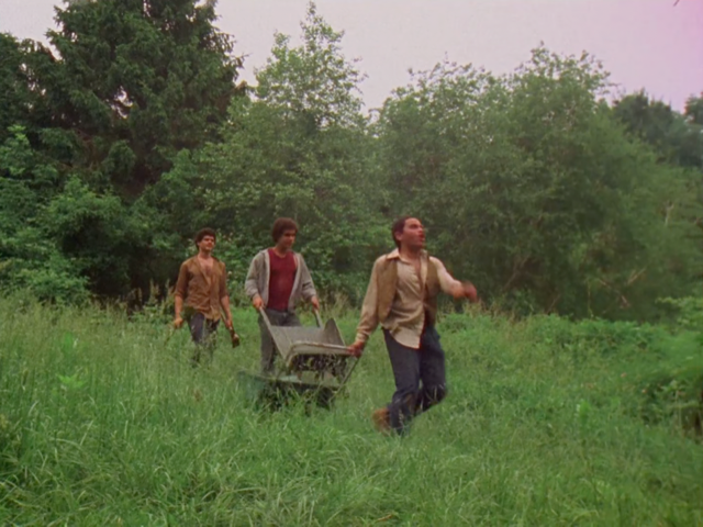 In a color film still, three young men walk in a line across an overgrown field with trees in the background. The middle one is pushing a wheel barrow.