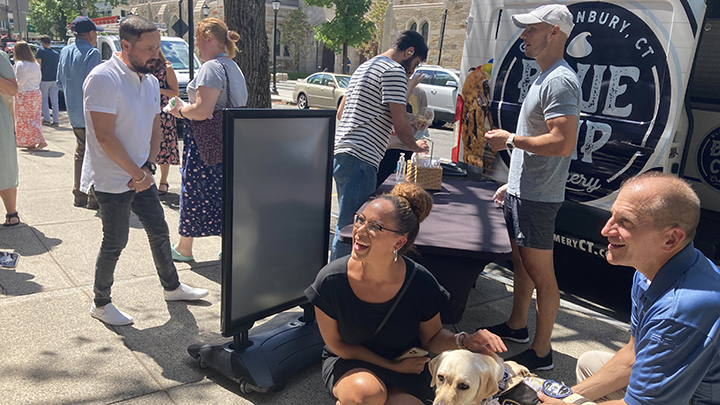 In the background, people gather around the ice cream table. In the foreground, a woman with her hair in a top bun, black dress, and glasses is laughing, while petting the service dog. To the right, the dog's handler in Yale police uniform smiles.