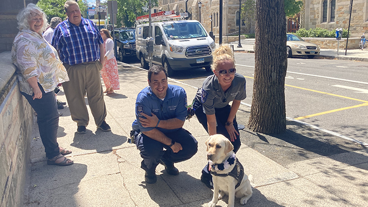 Two security guards pose with Heidi the labrador service dog. Both are crouching down and smiling. The man has black hair and the woman is blonde with sunglasses. To the right, a woman with white hair, a pastel-patterned shirt and jeans, and a man in a plaid shirt and long khaki pants look on smiling.