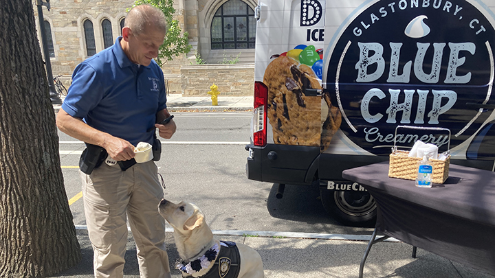 The service dog's handler stands above Heidi the dog holding a cup of ice cream. The dog points his nose up toward the cup. Rich, the handler, wears a blue short-sleeve Yale police shirt and khaki pants. The ice cream truck and the serving table are visible at right.