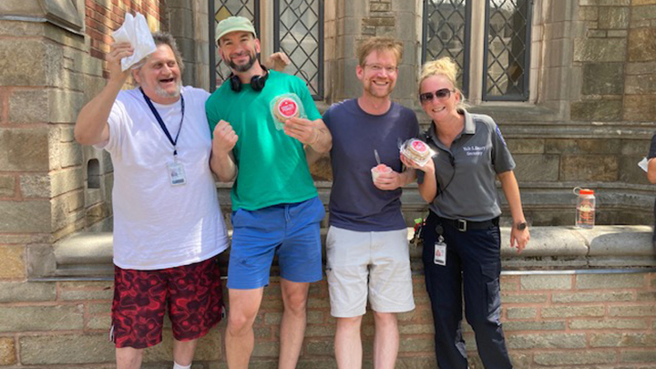 Four friends pose for the camera, holding up their ice cream sandwiches. The three men are wearing shorts. The blonde woman wearing sunglasses is a security officer in uniform. 