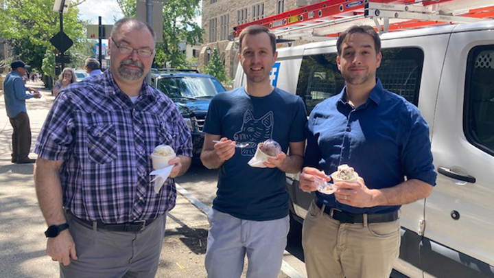 Three men pose for the camera, holding cups of ice cream. The man at left wears glasses and a van dyke and a blue and purple plaid short-sleeve shirt. The younger men with him both wear dark blue shirts and light-colored pants.