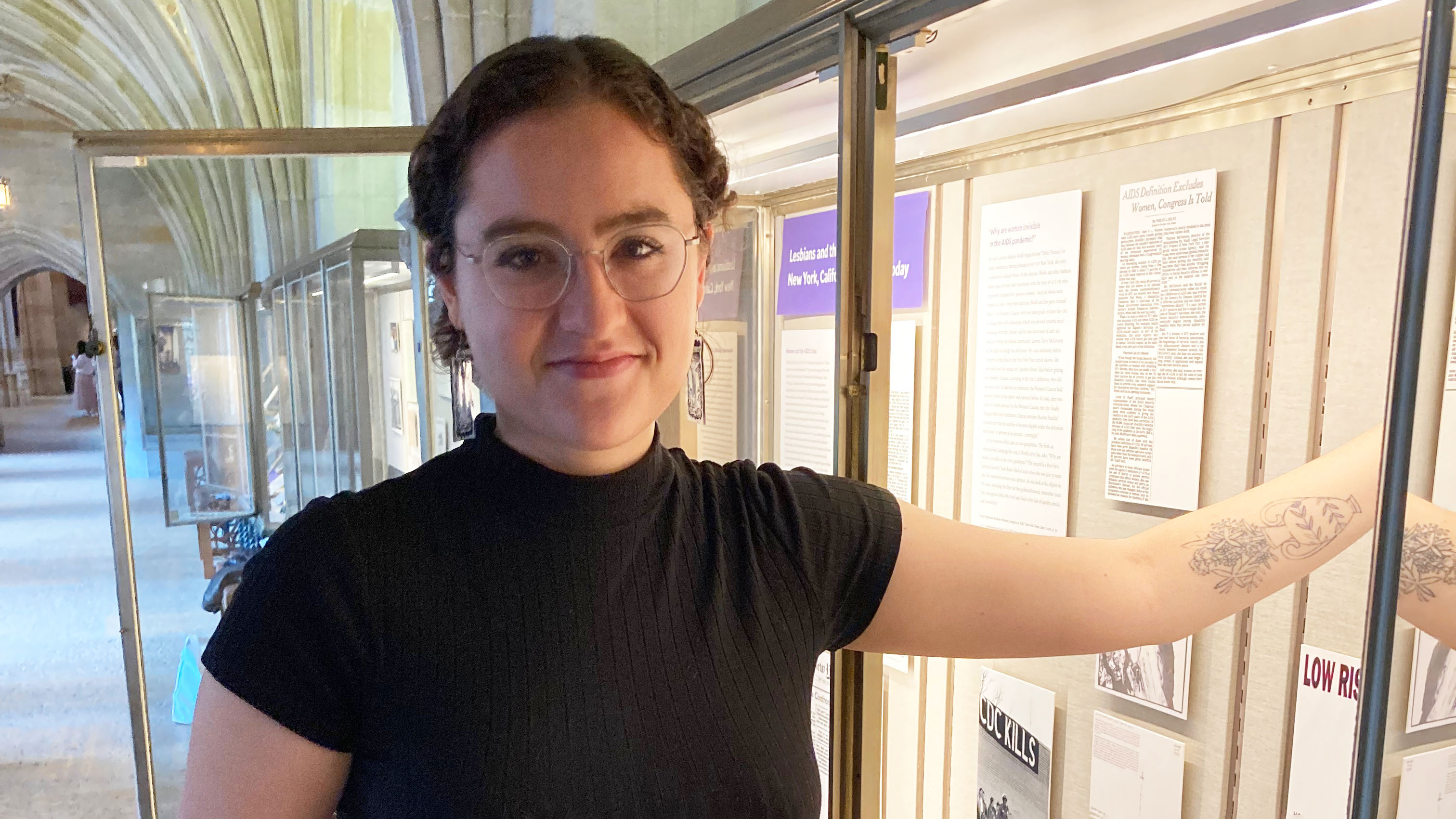 Curator Gabby Colangelo stands in front of open display case in Exhibition Corridor. She wears a short-sleeve black top, wire-rim glasses and long rectangular earrings. Her brown curly hair is tied back. She has a tattoo of a Greek vase with flowers on her forearm and reaches into the case, looking at the camera.