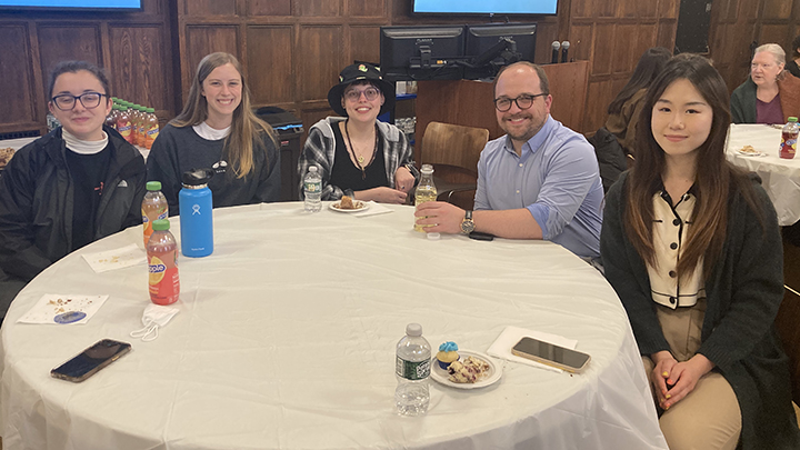 Five students--four women and one man--sit at round table. Two of the women and the man wear glasses. Drinks and plates of cookies and cell phones are on the table in front of them.