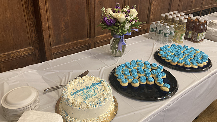 Table with white-frosted cake with blue frosting lettering that reads "Congratulations Class of 2023". Two plates of cookies with swirls of blue icing sit on black plates further down table. There are five rows of water and bottled drinks. A floral arrangement with white roses, blue flowers and a purple bow around the glass vase is at center of the table.
