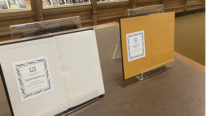Two open books, one with white end papers and one with brown end papers, are propped open in mylar stands on a tabletop, displaying decorative book plates on left pages, each shows a drawing of an open book at center and the words "In honor of" below, with a student's name written in blue script with graduation year "'22" below