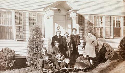 Black and white group photo of three women and a man standing in front of the front door of a clapboard house, a group of children in front, all wearing winter coats, ca. 1941