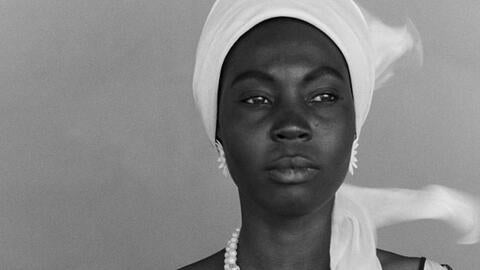 Black woman wearing white turban, large white flower earrings and white pearl necklace against a grey background