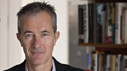 Portrait of Geoff Dyer in blue/white-striped button-down shirt and dark suit jacket, standing in front of wall of bookcases