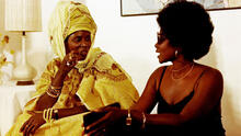 Two Senegalese women sit and talk indoors. One wears bright yellow traditional dress and headdress; the other wears a black tank top, hoop earrings, and sunglasses
