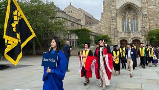 parade of people with caps and black robes and yellow stoles passing in front of stone library. Woman in blue carries banner and sign that reads "Ezra Stiles"
