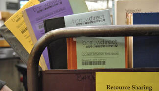 close-up detail of a metal library card holding four different colored books fanned out to show the BorrowDirect tracking labels on them. A sign on the cart reads "Resource Sharing"
