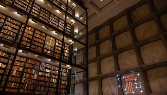 Interior view of Beinecke library of many stories of books on shelves behind glass across from marbled wall with projection that reads "Beinecke Celebrates 60"