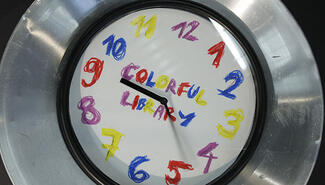 Clock face with wide silver rim with numbers with different-colored crayons and "Colorful Library" written in colorful crayons at center