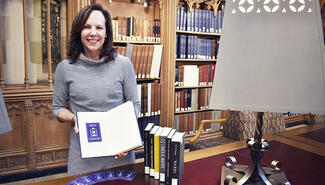 Women with shoulder-length brown hair stands in front of wood book cases holding an open book that has blue and white book plate inside cover. Five books and a grey lamp with metal base are standing on the table next to her.