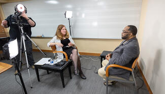 woman and man seated facing each other in a conference interview setting with videographer recording nearby