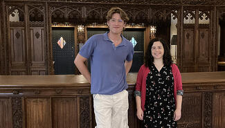 Tall young man with wavy brown hair, wearing wire-rimmed glasses, a blue shirt, white pants, and short necklace, stands next to small woman with dark hair, black and white flowered dress and red sweater. In the background are ornate wood-carved panels and they lean against a long, dark wood-carved surface that had once been the library's circulation desk..
