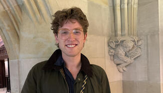 Young man with sandy brown hair and large amber-framed glasses, waring a dark shirt and jacket, smiles broadly and poses in front of carved column in the exhibition corridor of Sterling Memorial. The small carved figure is of a crouched man reading a book, one page of which has the carved word "Joke." 
