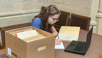 Woman writes at a desk beside a box of manuscripts