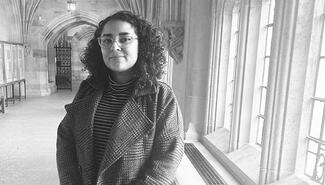 Woman with curly dark hair and wire-rimmed glasses poses alongside windows, with hallway and arched entrance behind her. The photo is black and white. She wears a striped turtleneck and plaid coat.