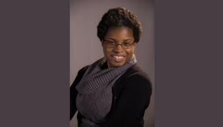 Black woman with glasses ad purple cowl neck sweater and black cardigan smiles at camera