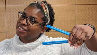 Smiling Black woman wearng white turtleneck sweater, glasses, and colorful hairband, holding two blue pencils printed with "catalog of US Government Publications" in white