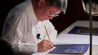 man in white tunic with glasses bends over blue paper drawing with bamboo pen