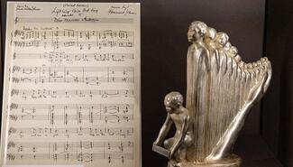 Hand-written sheet music titled "Lift Ev'ry Voice And Sing"; at right a small metal sculpture of a harp made up of standing figures with crouching boy at front