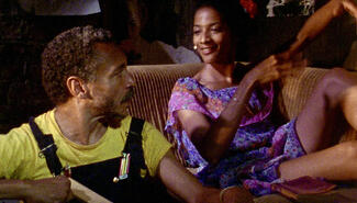 Film still of woman in purple print dress sitting on a couch, smiling at a man with in yellow T-shirt and overalls sitting next to her.