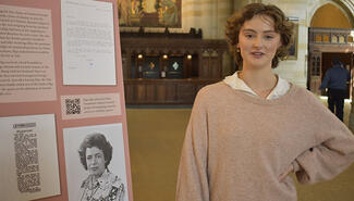 Young woman with short curly brown hair wearing pink sweater and white shirt stands with hand on hip next to display with photo of Elsa Wasserman and copies of documents